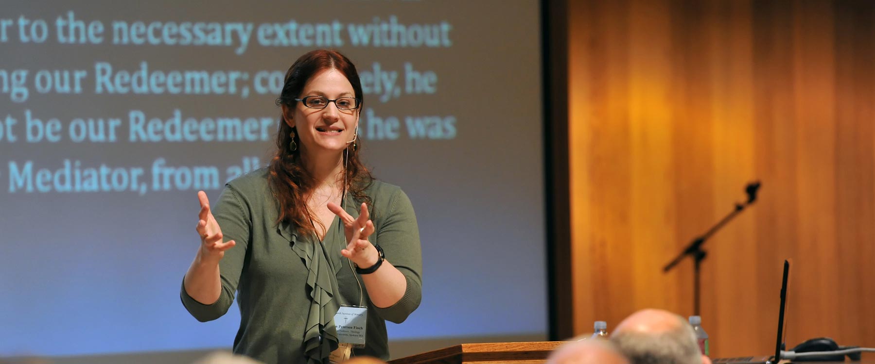 A speaker wearing a name tag and microphone headset, speaks in front of a projector screen.