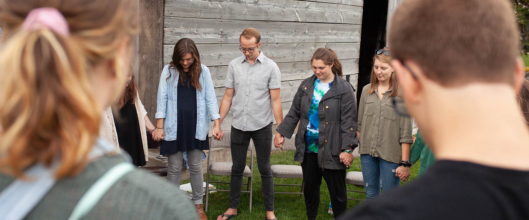 A circle of students hold hands, bow their heads and pray together outdoors.