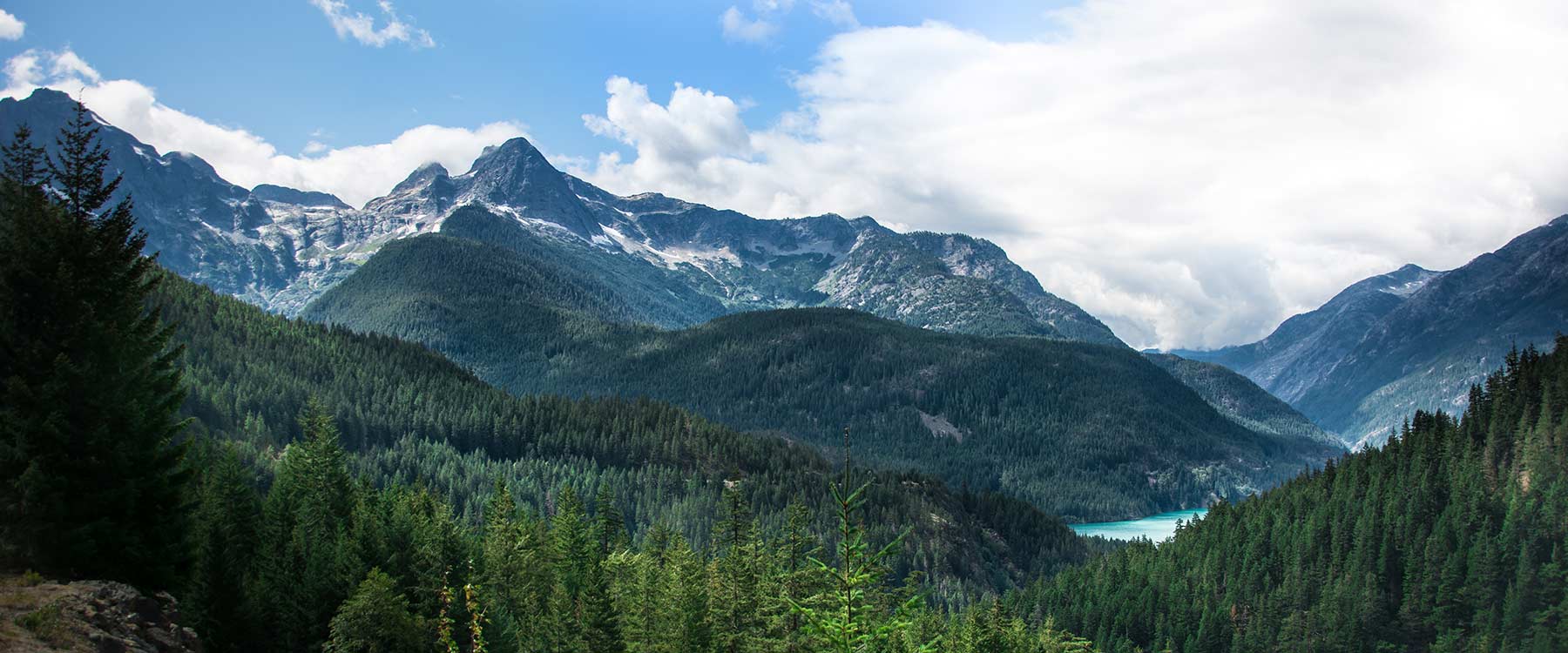 Scenic view of lush green forest and mountains beneath a partly cloudy sky, with a turquoise lake peeking through the hills.