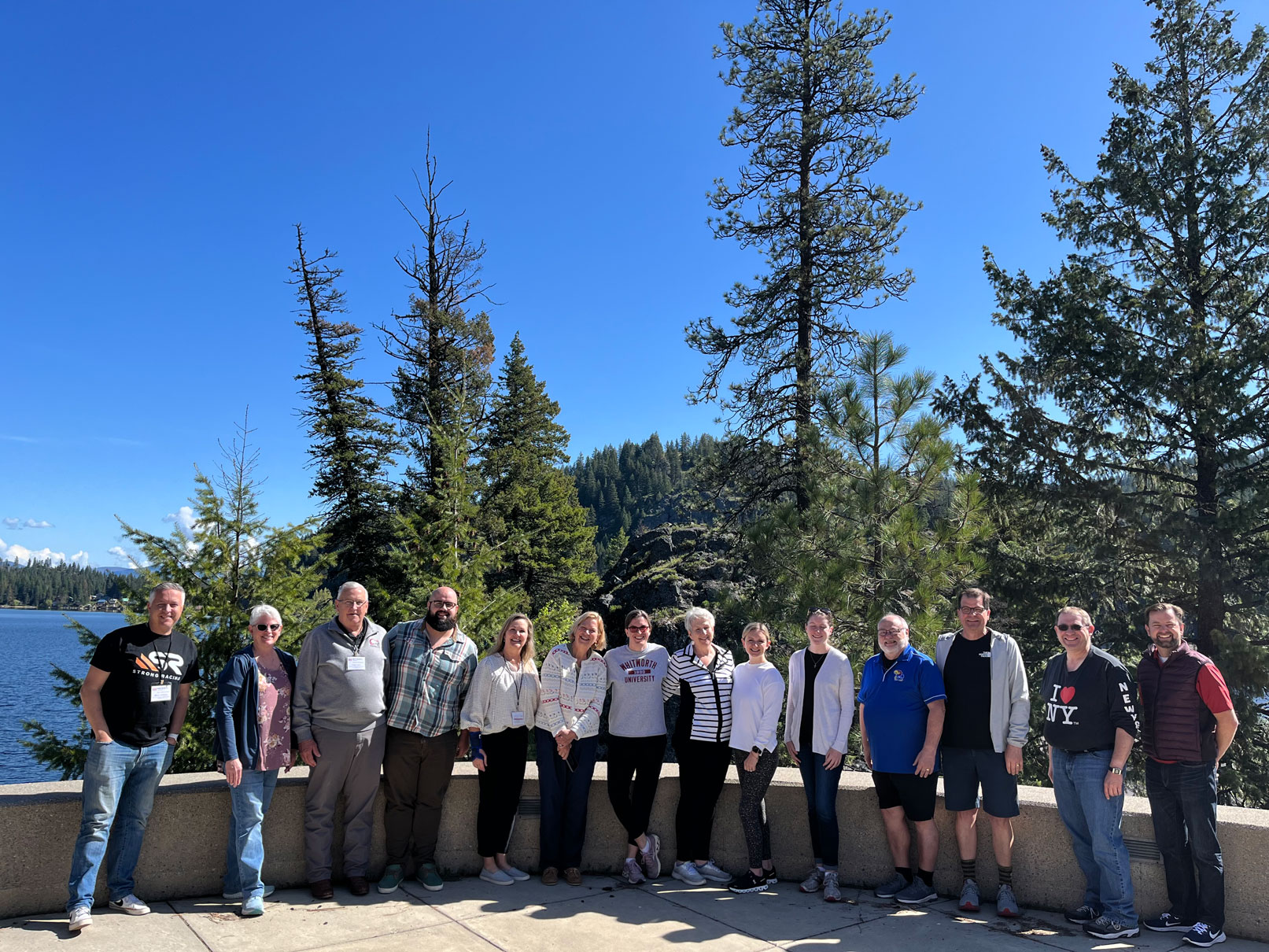 A group of adults poses in front of a lake and trees under a clear blue sky on a sunny day.