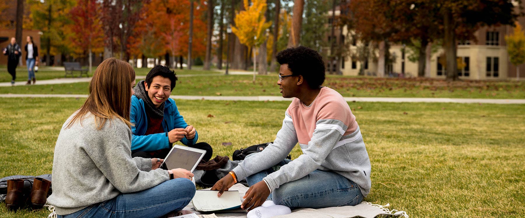 Three students dressed in fall clothing sit on the grass with notebooks, engaged in conversation.