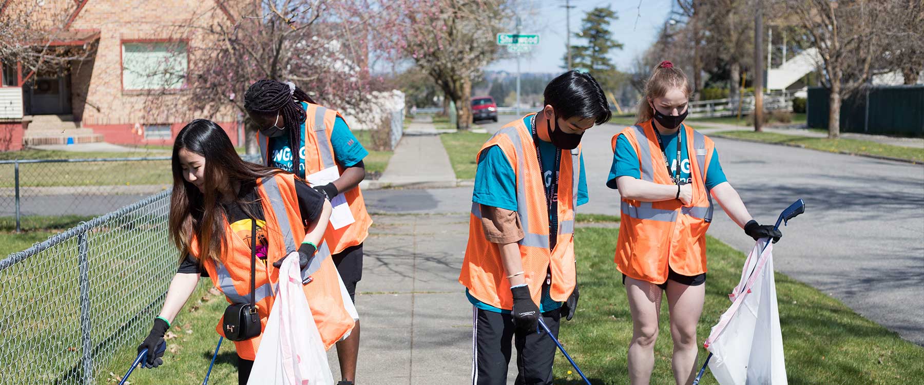 Four student volunteers wearing orange vests and gloves pick up trash along a residential street..