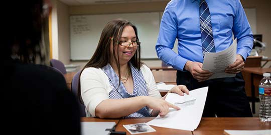 An adult student sits at a desk and discusses a paper with a professor who stands nearby.