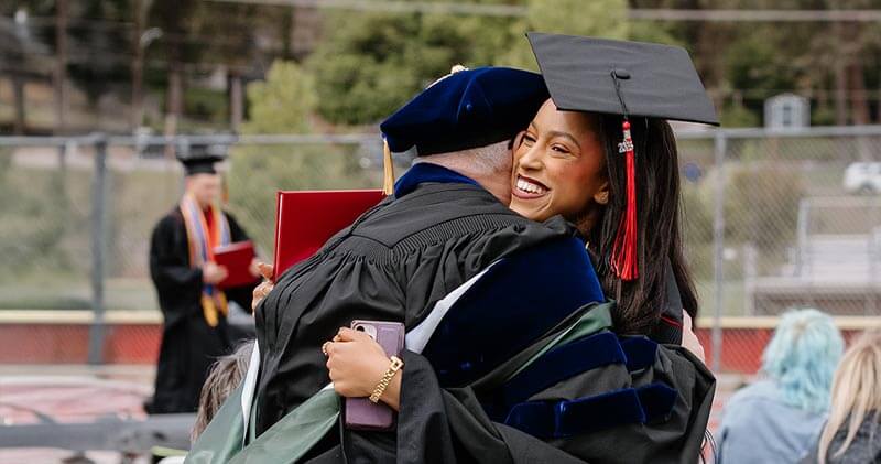 A professor and student in graduation regalia embrace joyfully at an outdoor commencement ceremony.