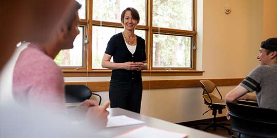 A professor stands in a classroom near a large window, smiling and listening to a student.