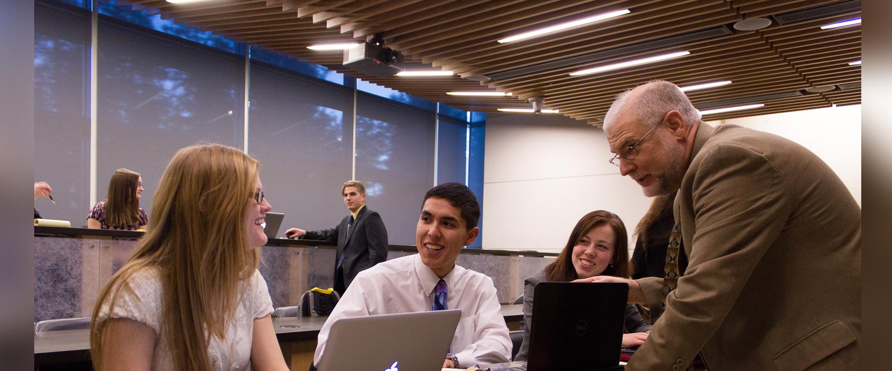 A professor leans over a shared computer desk and engages with three students, all of whom are smiling.