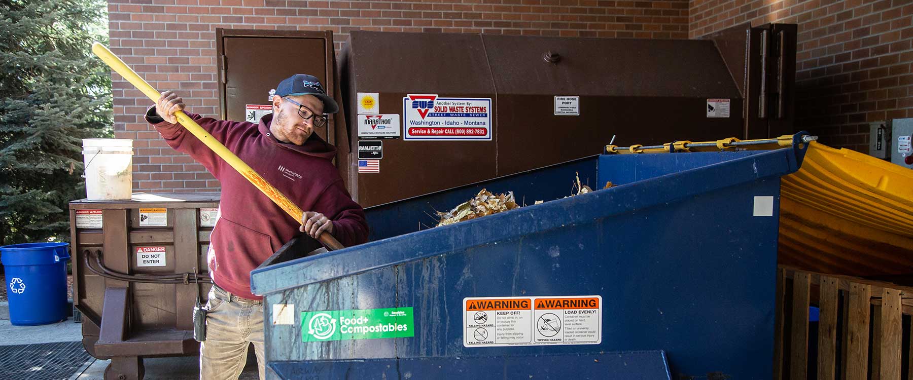 A facilities services staff member uses a shovel to rotate the contents of a compost bin.