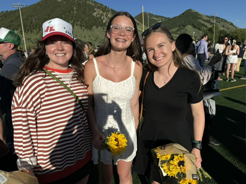 A young graduate stands flanked by two supportive friends outdoors on a sunny day.