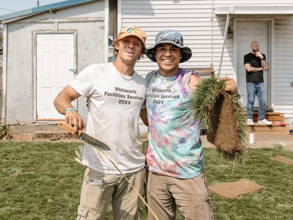 Two people smile in the front yard of a white house, one holds a large piece of sod, and one holds a garden tool.