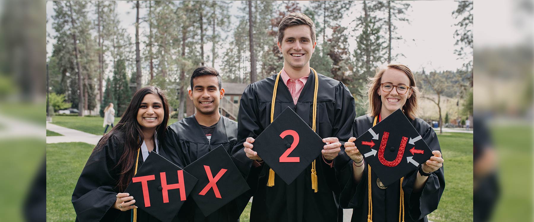 Four students stand in a row wearing graduation robes and smiling. The top of their caps say, 
