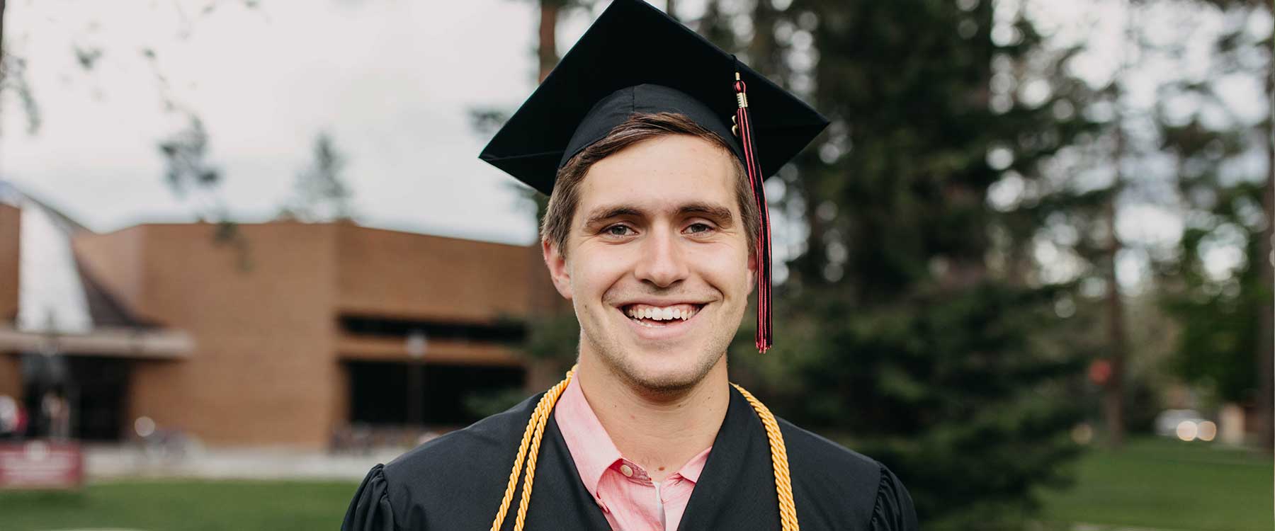 Micah stands in front of the HUB wearing his graduation robes and cap. He smiles at the camera.