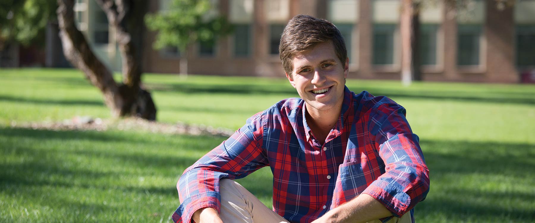 Micah sits on the grass in the middle of campus. He is posed, smiling for the camera.