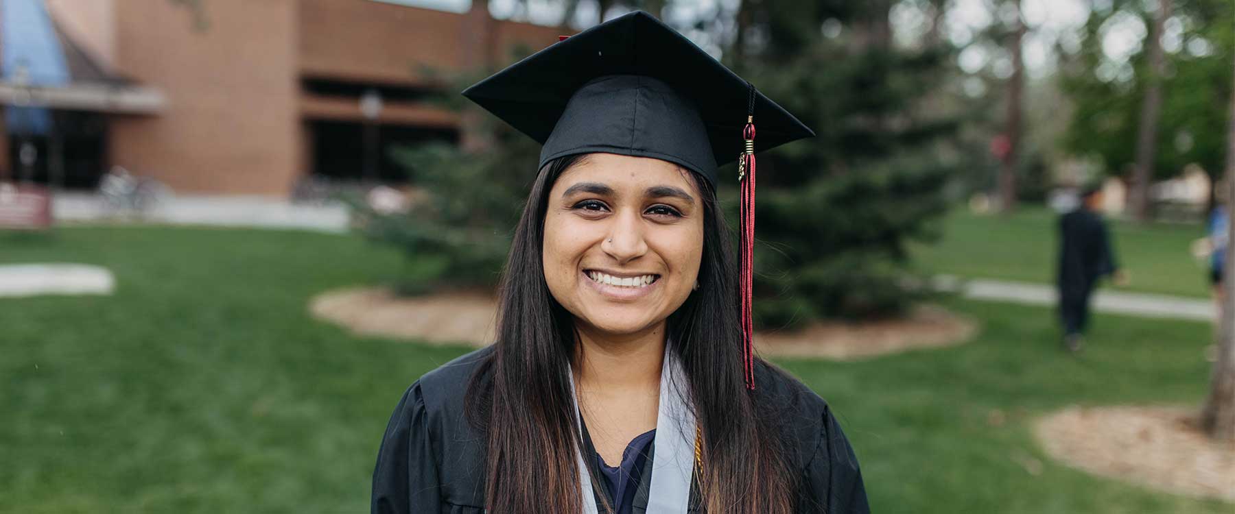 Sarah stands on the grass outside of the HUB wearing her graduation cap and gown. She smiles at the camera.