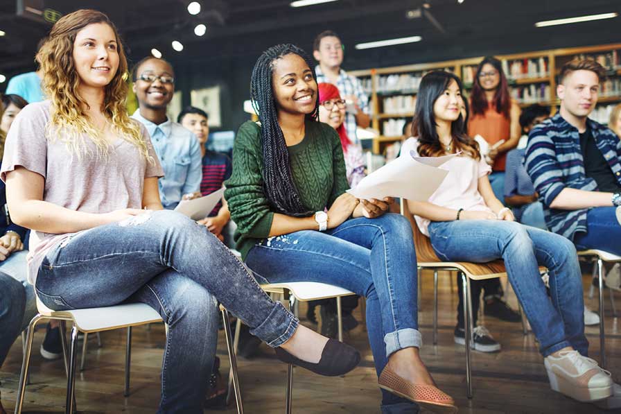 A diverse group of casually dressed students sit in a classroom, attentively looking forward and holding papers.