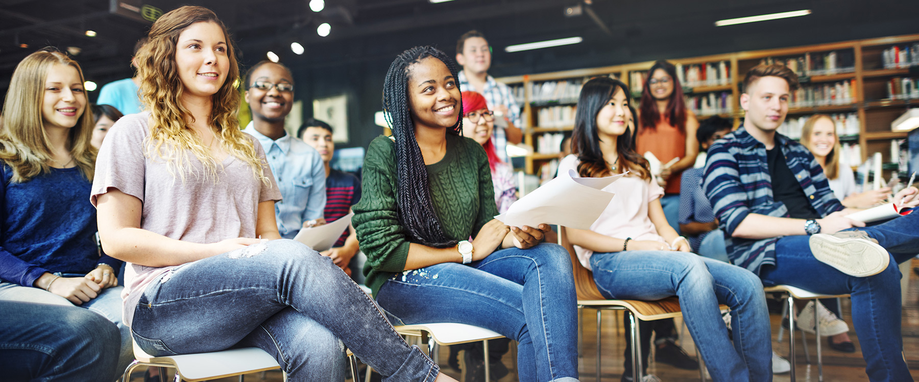 A diverse group of casually dressed students sit in a classroom, attentively looking forward and holding papers.