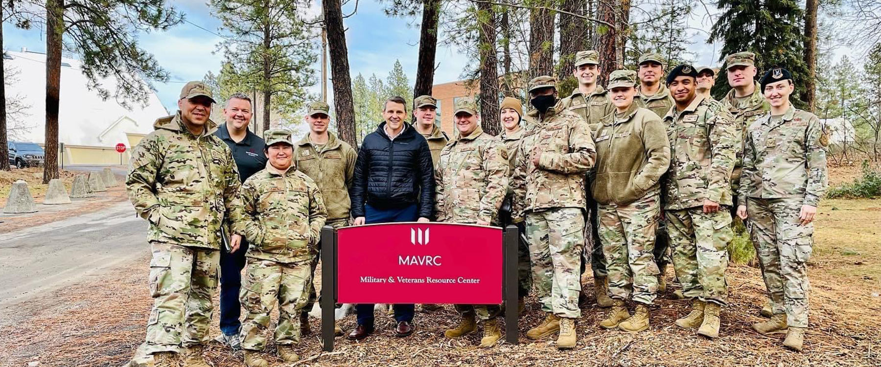Group of people in military uniforms and casual clothes stand by MAVRC sign in a wooded area.