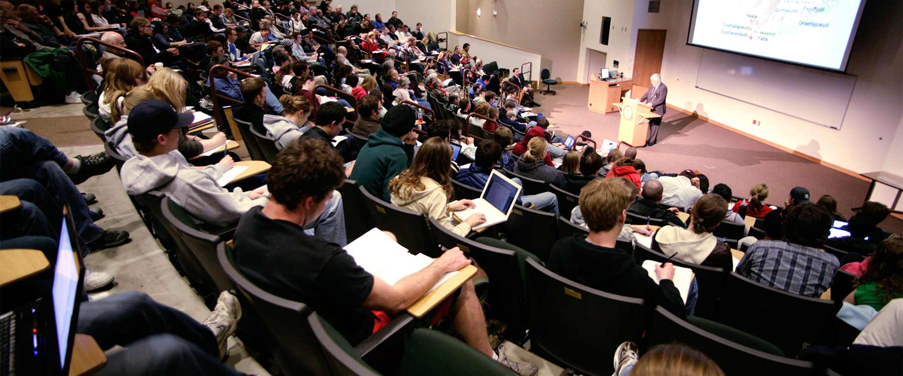 Students sit in a tiered lecture hall and take notes. At the front, a professor speaks.