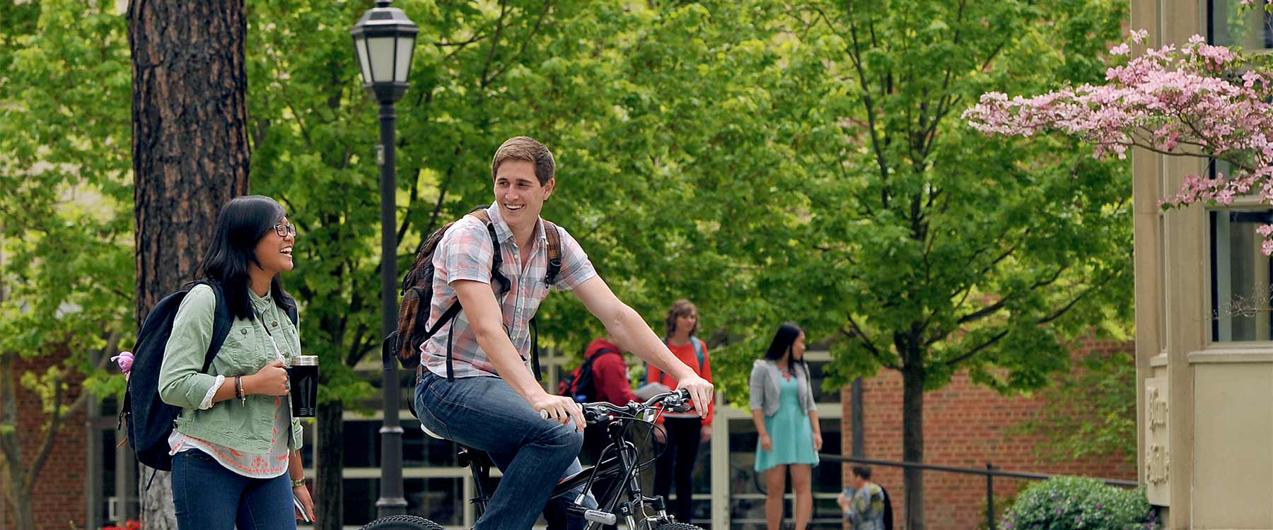 A student rides his bike next to another student who is walking. They look at each other and laugh.