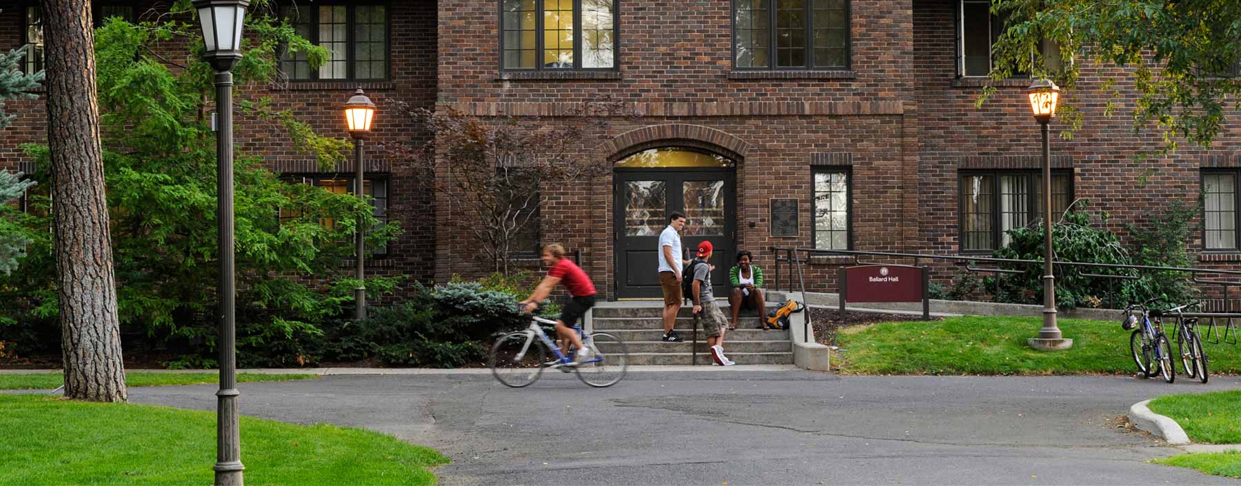 A student passes by on a bicycle as two students stand and one sits on the steps of a stately brick building at dusk.