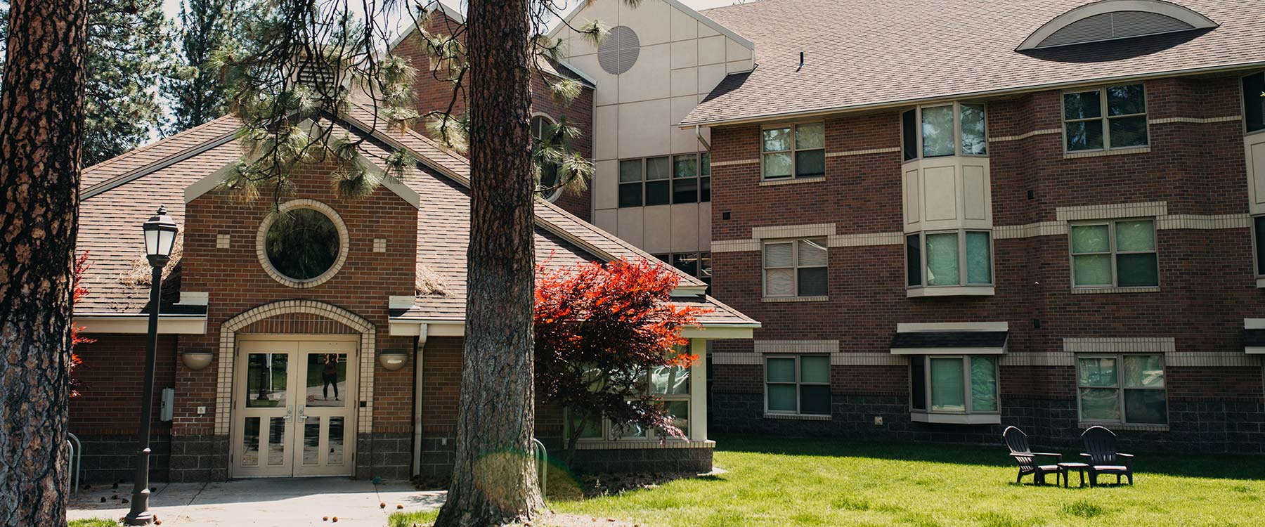 A multi-story brick building with arched windows and a lawn, surrounded by tall trees and outdoor seating.