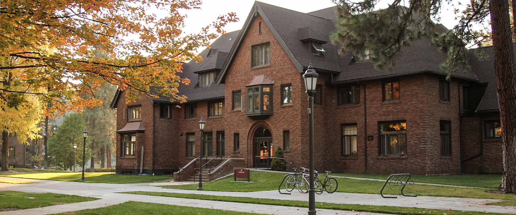 A brick university building with a gabled roof, surrounded by trees with autumn foliage, and a bike rack in front.