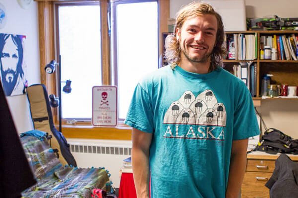 A student stands and smiles in his dorm room. Behind him is a cluttered bookshelf, skis and a futon.