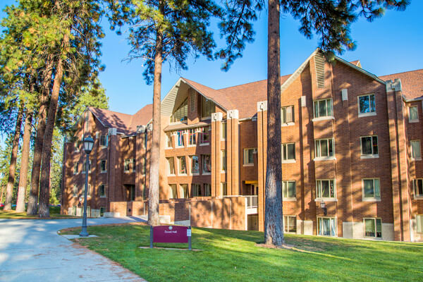 A four-story building with multiple gabled peaks stands among tall pine trees on a clear day.