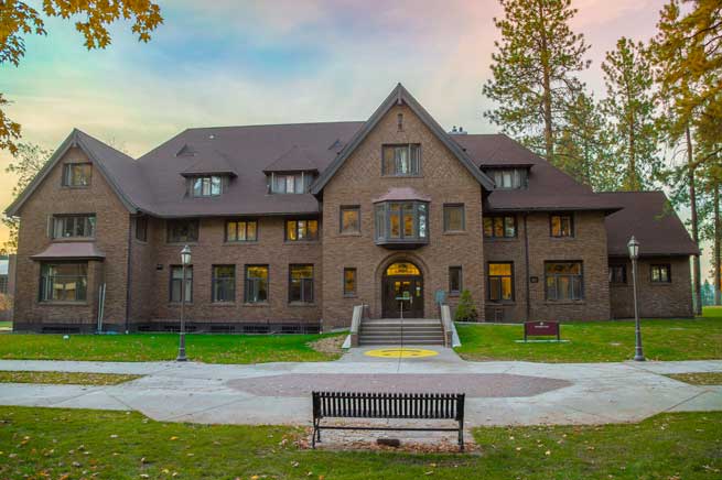 A large brick building with a steep roof, surrounded by trees and a park bench in the pastel light at sunset.