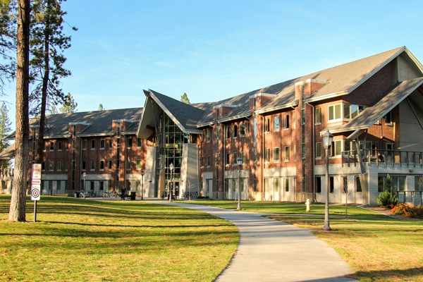 Dapple sunlight hits Oliver Hall, a four-story brick building, through tall pine trees.