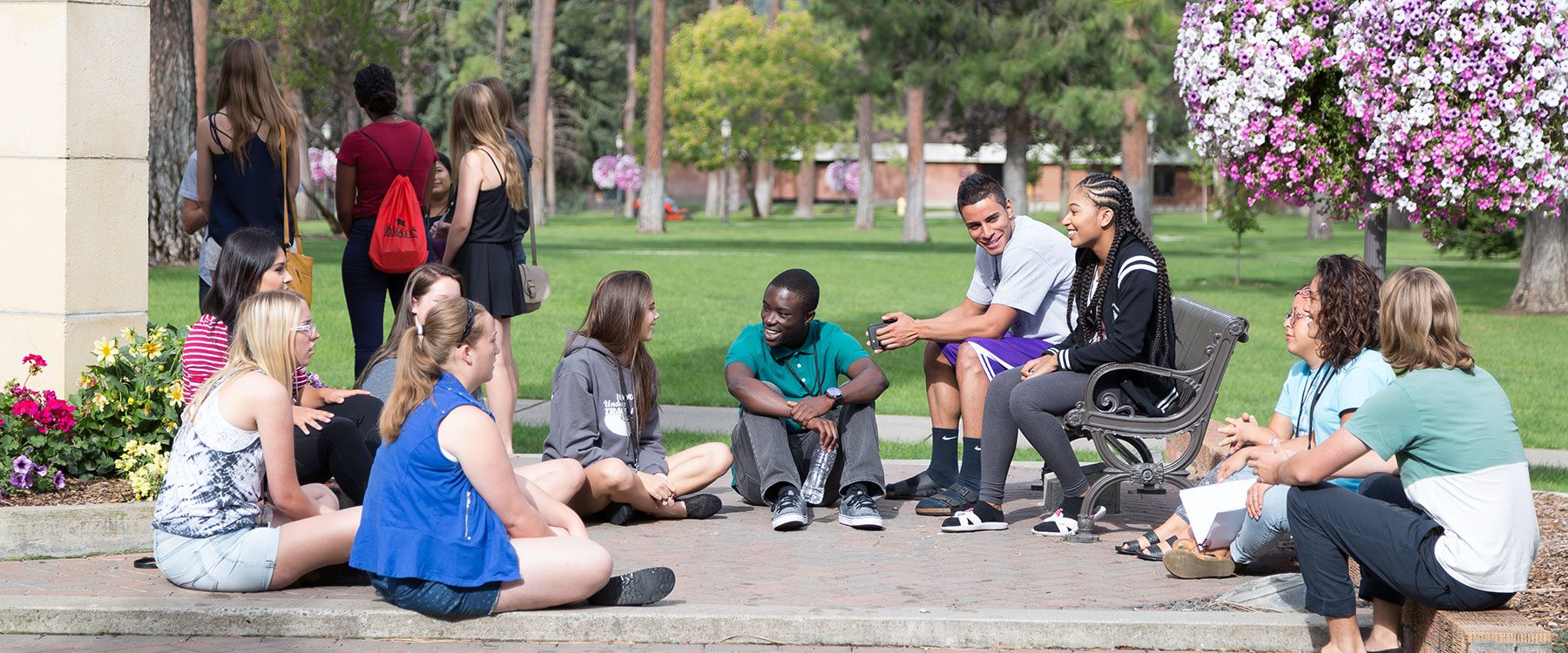 A diverse group of students sit in a circle somewhere on campus. They smile and talk.