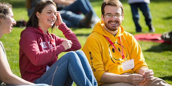 Close-up of three students sitting on the grass at an outdoor event, laughing and enjoying themselves.