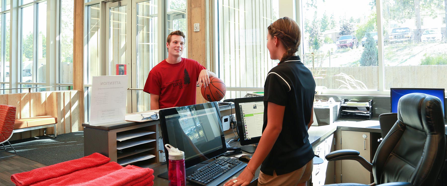 A student holding a basketball stands at the front desk and speaks with a U-Rec student employee.