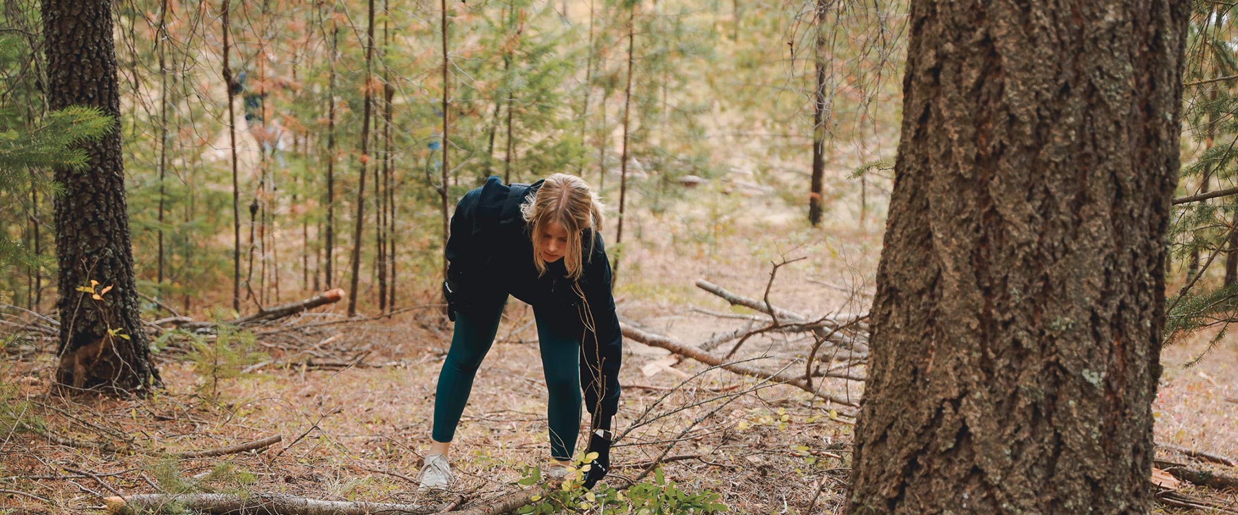 A student stoops over to pick up a branch off the trail in a forested area.