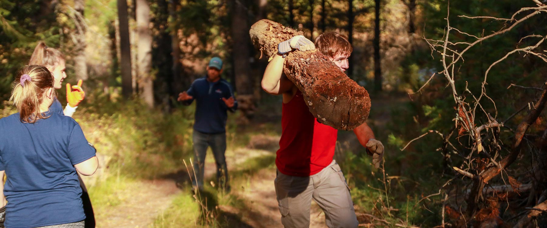 Students working together on a forest trail with one carrying a large log.