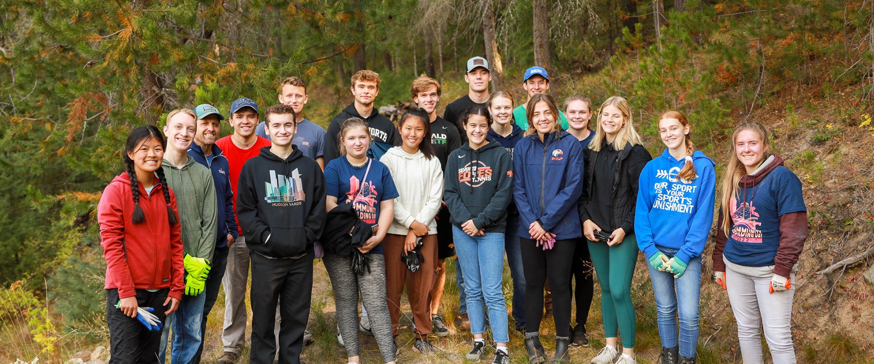 A diverse group of students, most wearing work gloves, pose for a picture in a wooded area.