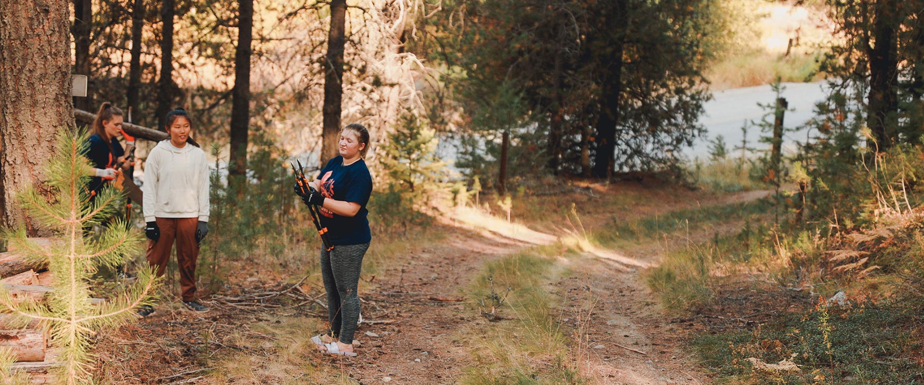 Three students in work clothes, holding tools, stand on a forest trail by a lake in autumn, surrounded by tall trees.