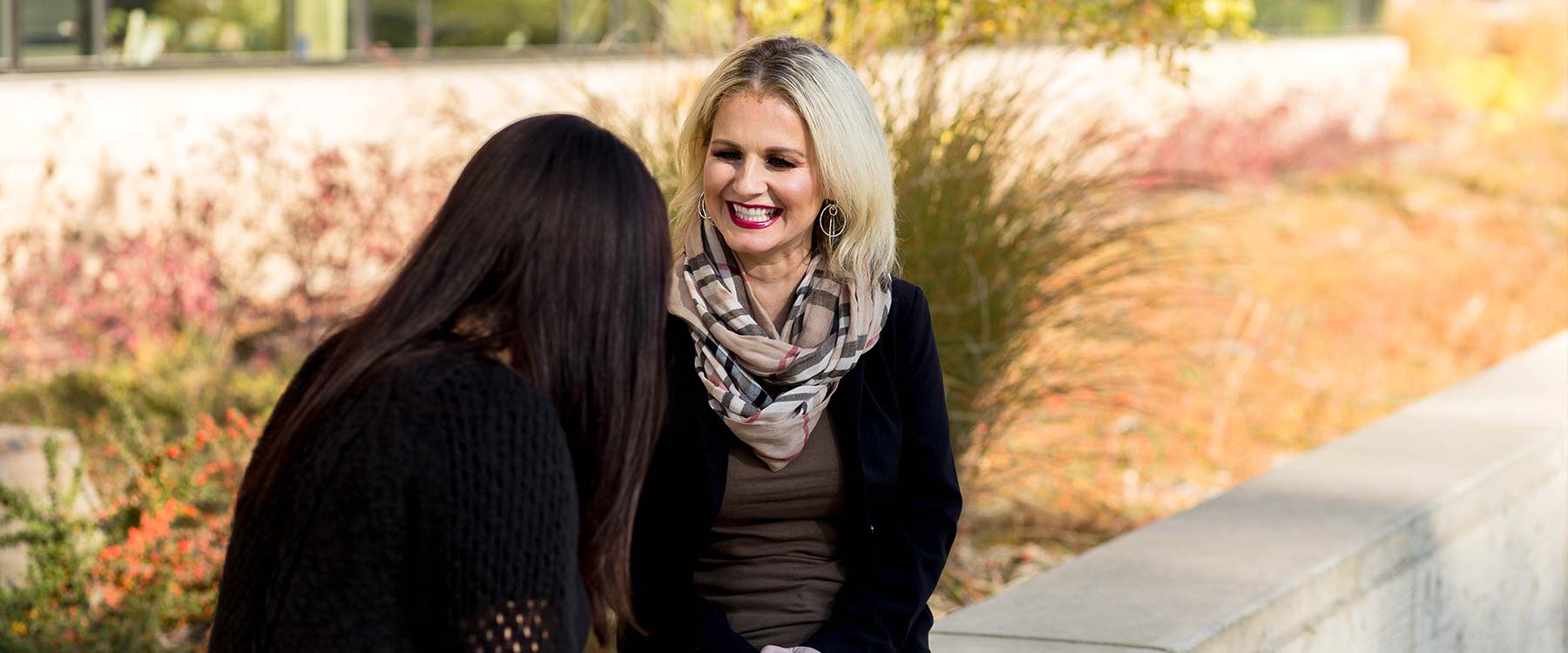 Two women sit and talk, smiling, outside on a sunny day, with autumn foliage in the background.