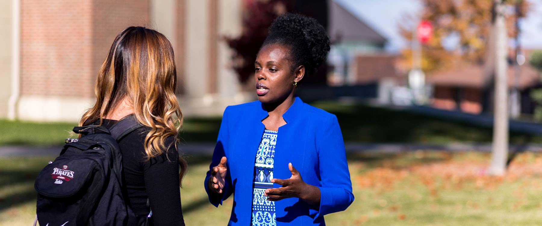 A professor and a student stand outside an academic building on a sunny day, engaged in deep conversation.