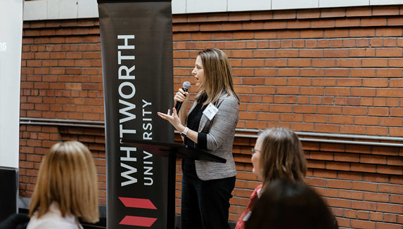 A woman speaks into a microphone at a podium near a 