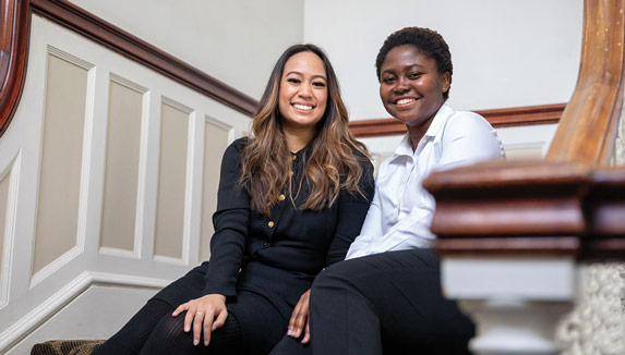 Two women sit together on an interior stairwell in a building with white paneled walls.