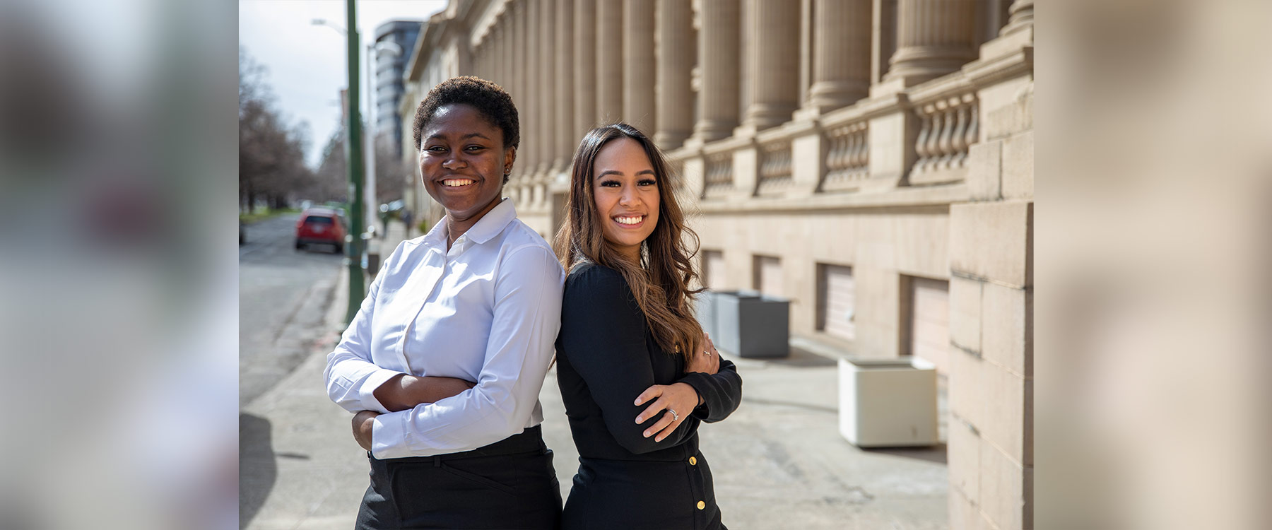 Two professional women stand back-to-back with arms crossed, both smiling confidently, near an urban building with columns.