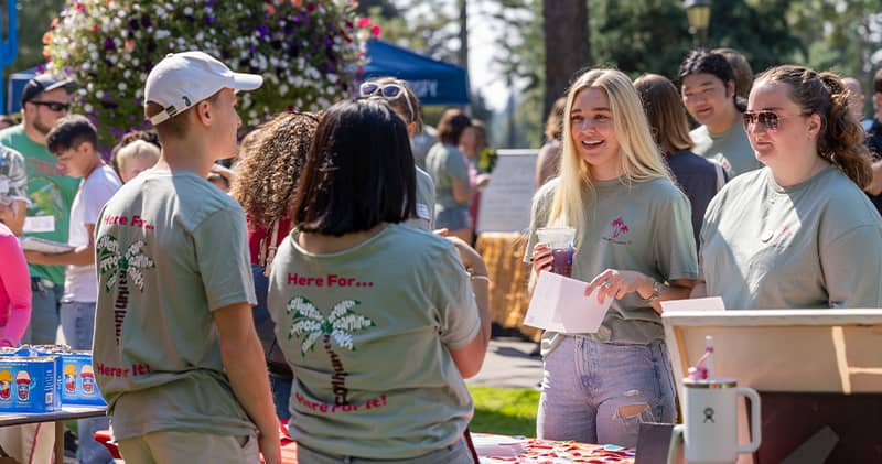 Multiple students converse cheerfully at a booth in a festive outdoor setting.