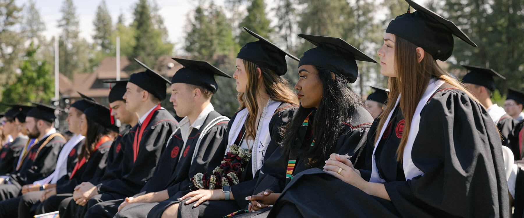 Graduates in caps and gowns sit in rows outdoors during a graduation ceremony, with trees in the background.