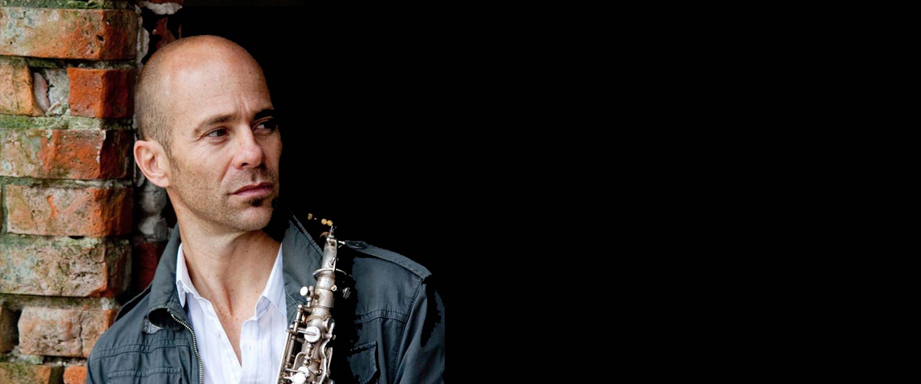 Wildy Zumwalt leans against a brick wall holding a Saxophone. He looks off to the right.