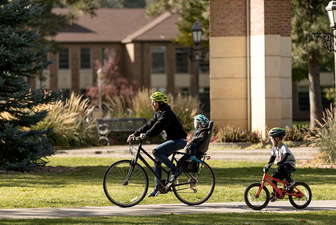 An adult and two children, all wearing helmets, ride bicycles on a sunny day with buildings and greenery in the background.