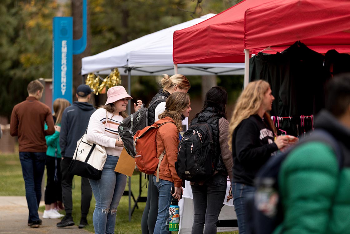 Multiple people standing in line and browsing at outdoor tents with merchandise.