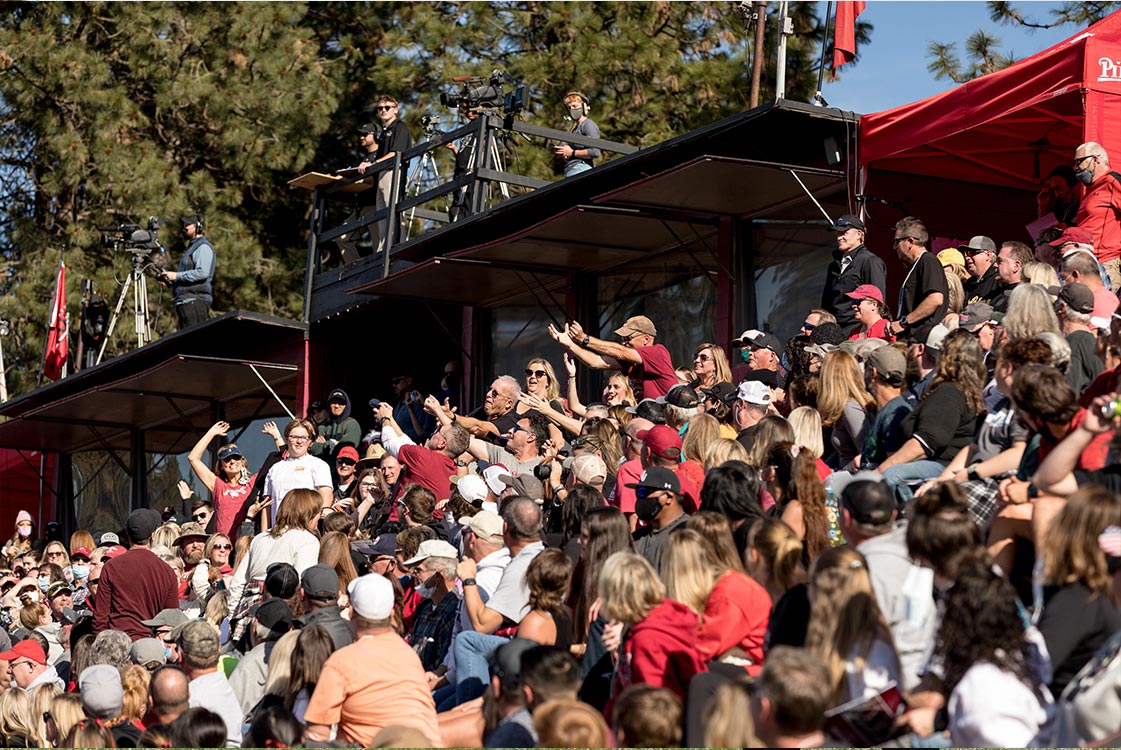 A large crowd of spectators in an outdoor setting, many wearing red, smiling and cheering.
