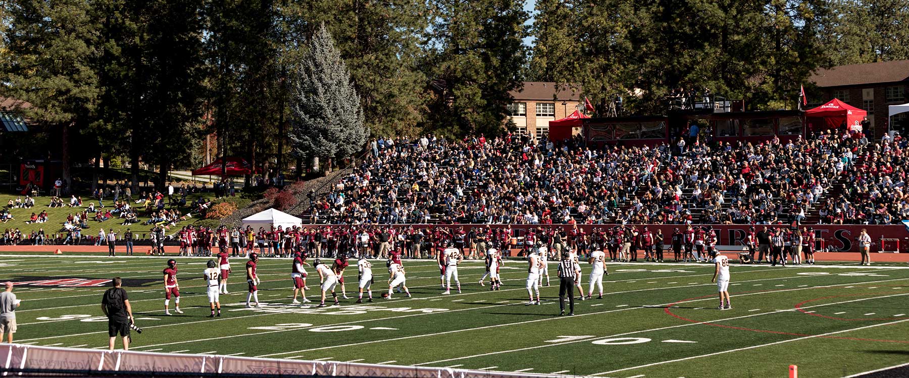 A football game in action with players on the field and a large, cheering crowd in the stadium on a sunny day.