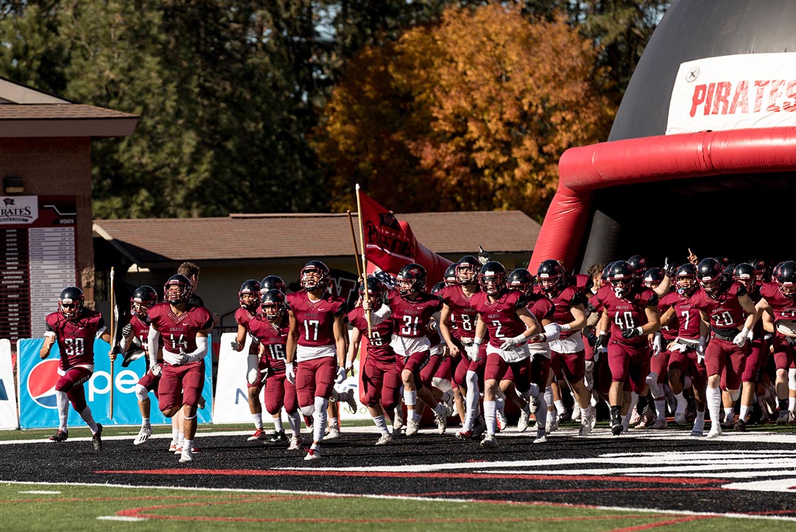 Football players in red uniforms run out of an inflatable helmet onto a field, one carrying a Whitworth Pirates flag.