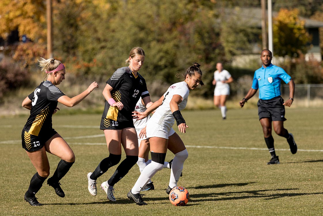 Three soccer players chase a soccer ball while a referee watches nearby.